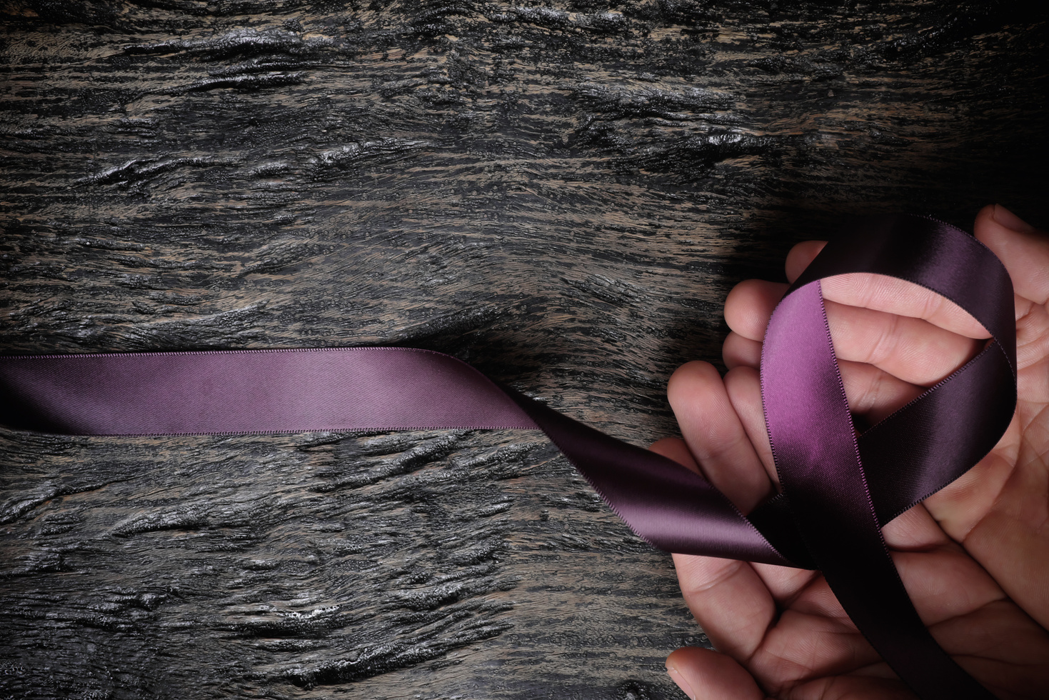 Top view of male hand holding purple ribbon on wood background with copy space. Pancreatic cancer, epilepsy, leiomyosarcoma, lupus, Alzheimer's disease and cystic fibrosis awareness concept.